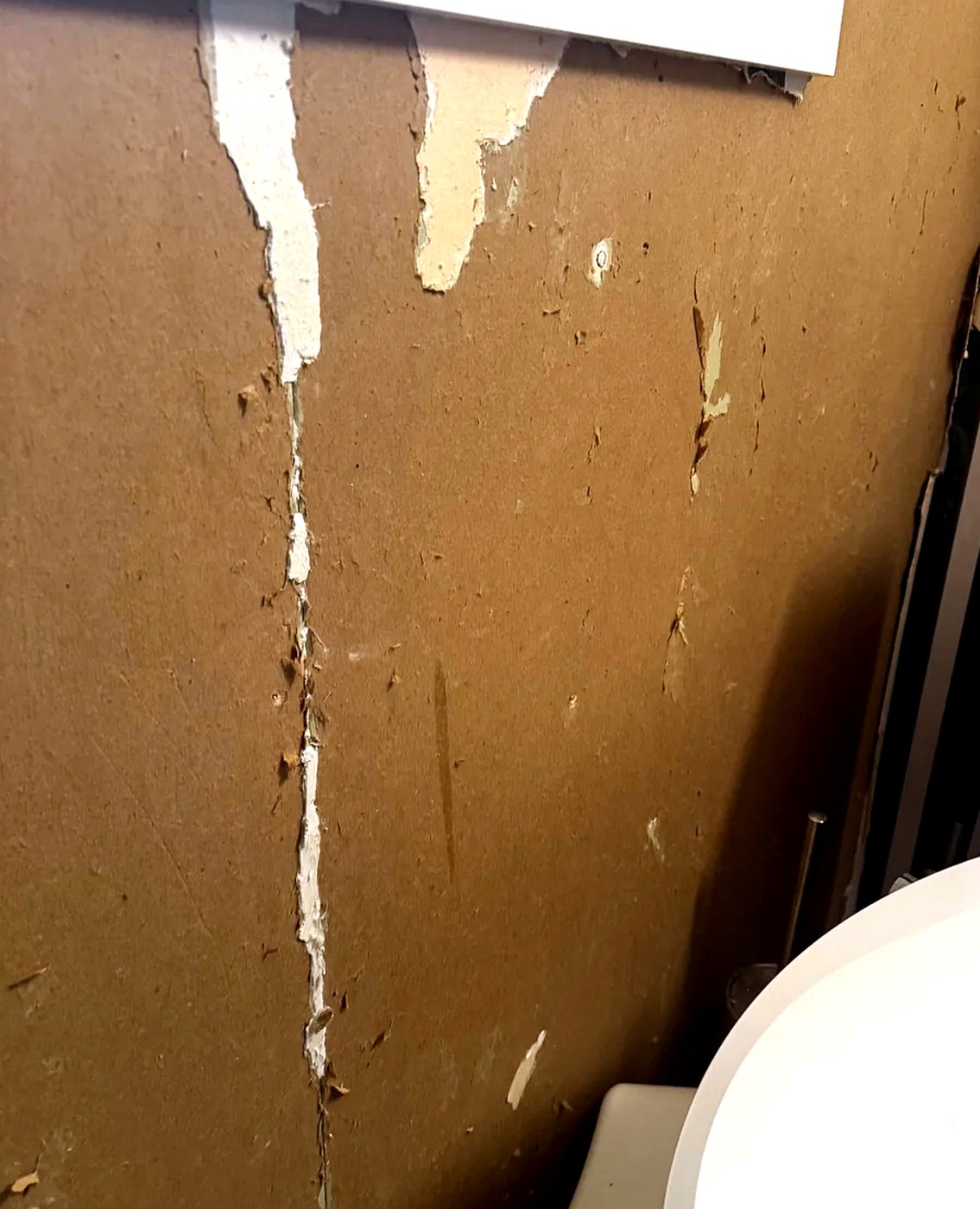 Bathroom wall with ripped drywall paper near a toilet and window trim